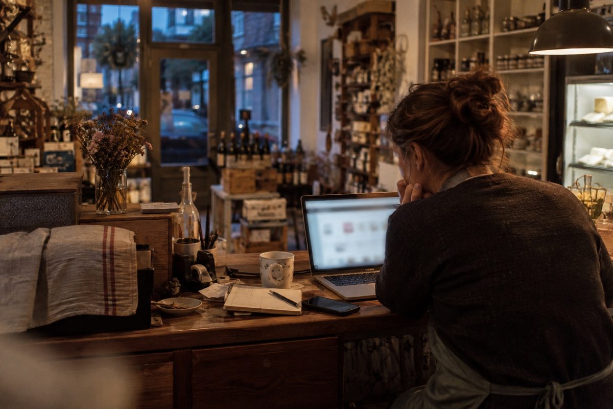 Eine Frau sitzt am Abend in einem kleinen Laden mit dem Laptop am Tresen, daneben liegen Notizbuch, Handy und eine Tasse.
